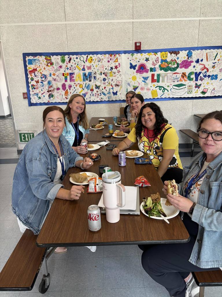 Staff members enjoy lunch together in a school cafeteria decorated with colorful student artwork and literacy-themed murals.