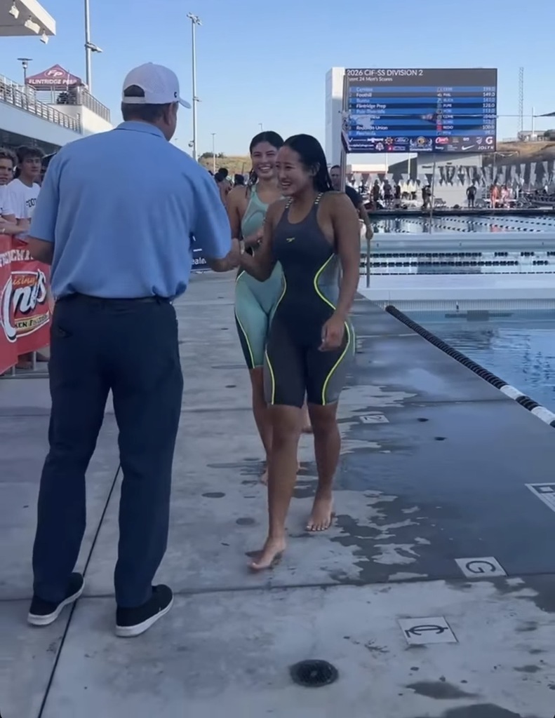 Swimmers in racing suits shake hands with an official beside an outdoor pool during a swim meet.