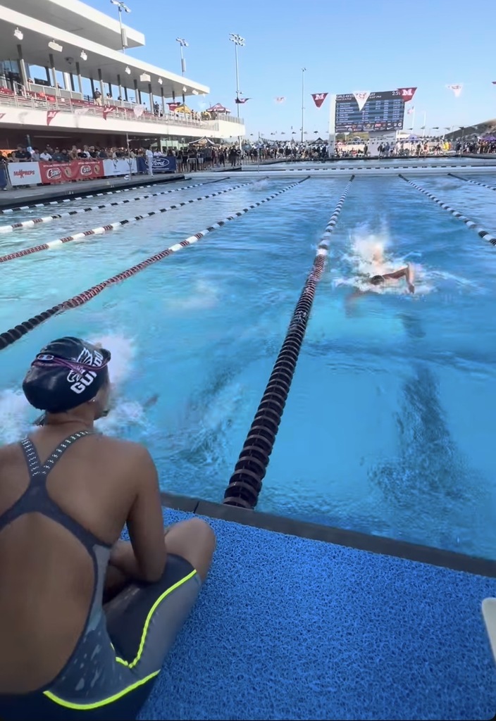Swimmer in a cap and suit watches a race in an outdoor pool with multiple lanes and spectators in the stands.