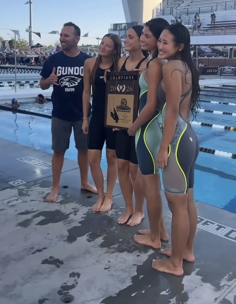 Four swimmers and their coach stand poolside holding a 2026 championship plaque at an outdoor swim meet.
