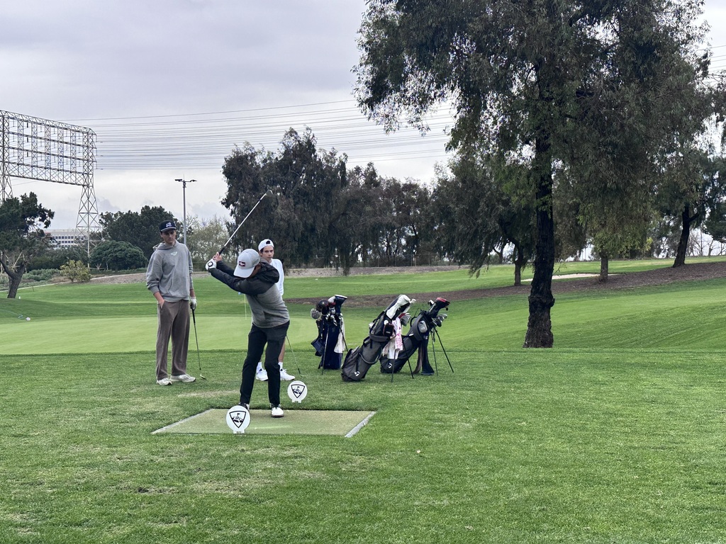 Student golfer swings from the tee box while teammates watch beside golf bags on a cloudy course.
