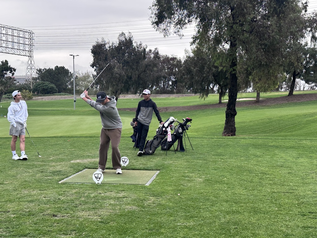 Student golfer prepares a drive on the tee box as teammates stand nearby on a green golf course.
