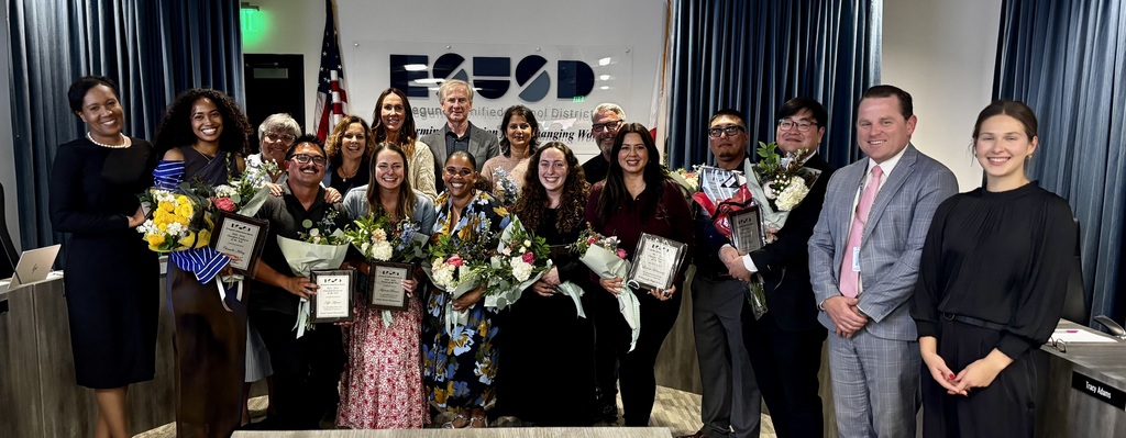 Group of diverse individuals holding plaques and bouquets, posing for a celebratory photo in front of a school district logo and American flag.