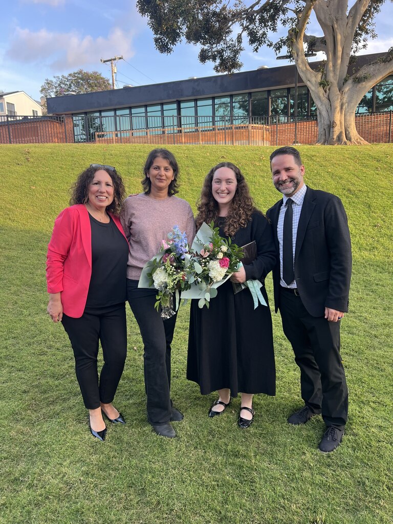 Four people standing on grass, one holding a bouquet, with a building and large tree in the background.