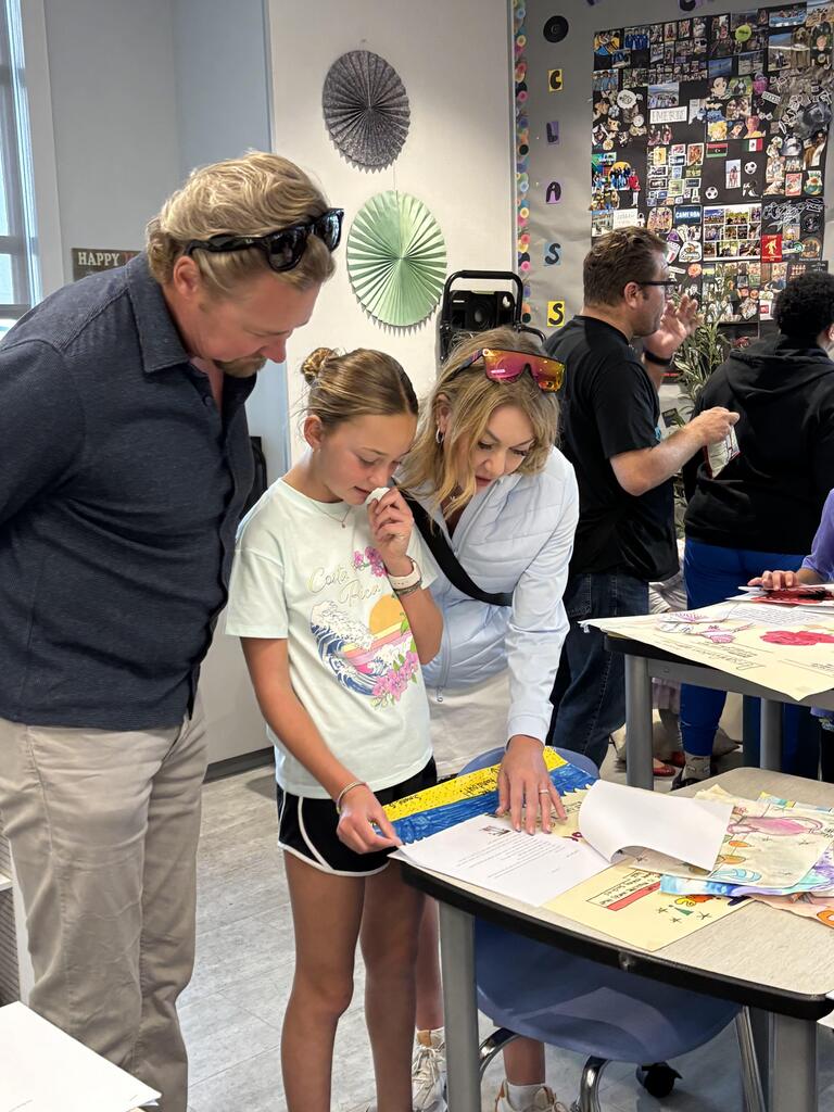 Parents speak with students beside a desk displaying schoolwork and artwork in a bright classroom.