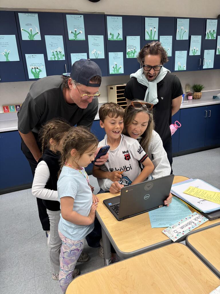 Parents and children gather around a laptop as a student shares classroom work during school open house.