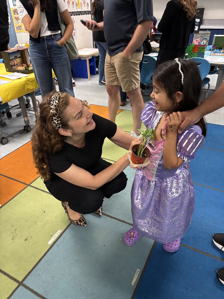 Teacher smiles while presenting a potted plant to a young student in a purple princess dress during classroom night.