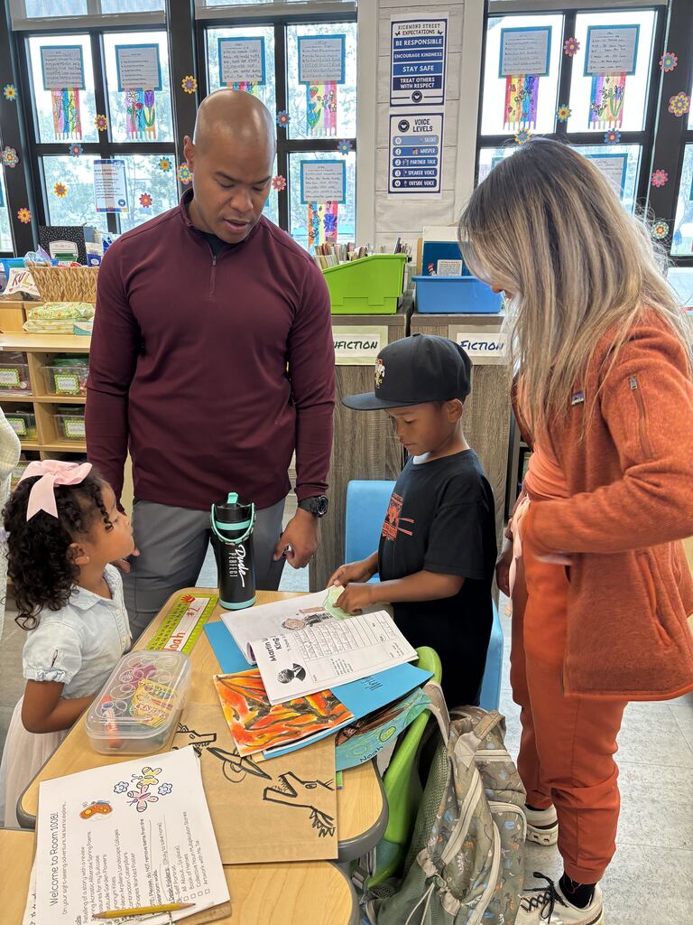 Parents speak with students beside a desk displaying schoolwork and artwork in a bright classroom.