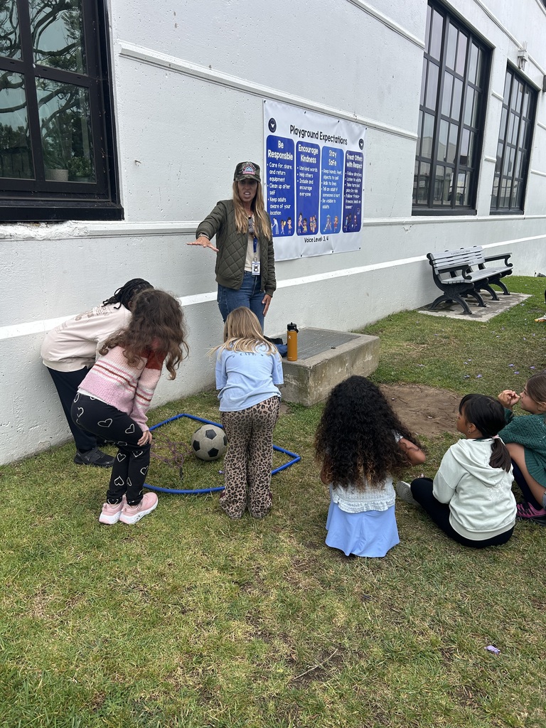 Teacher instructing a group of young children gathered on grass near a building with a soccer ball inside a blue hoop.