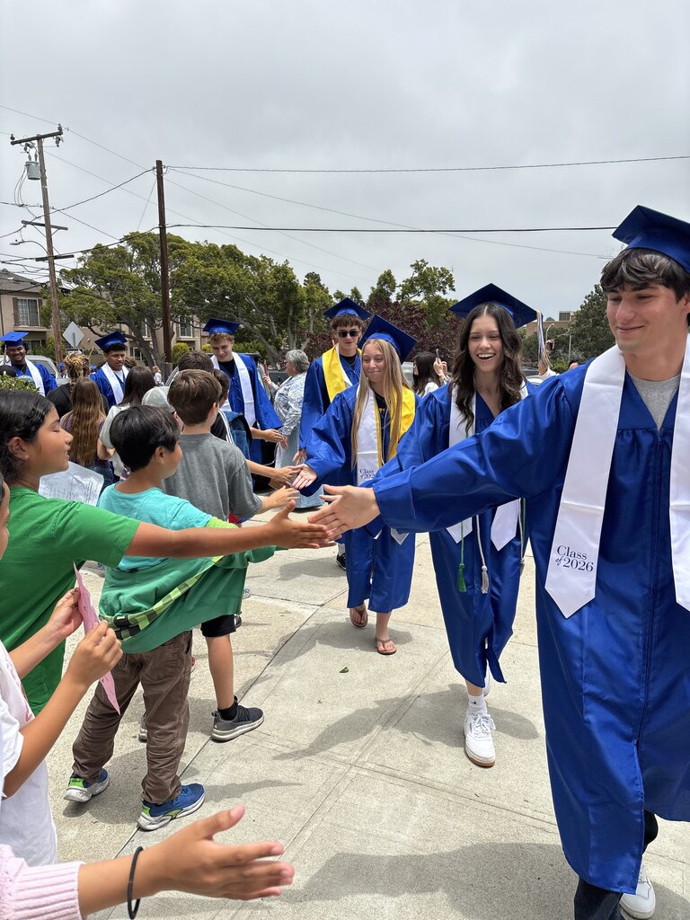 Graduating seniors in blue caps and gowns participate in a school walk, exchanging high-fives with younger students and families gathered outside.