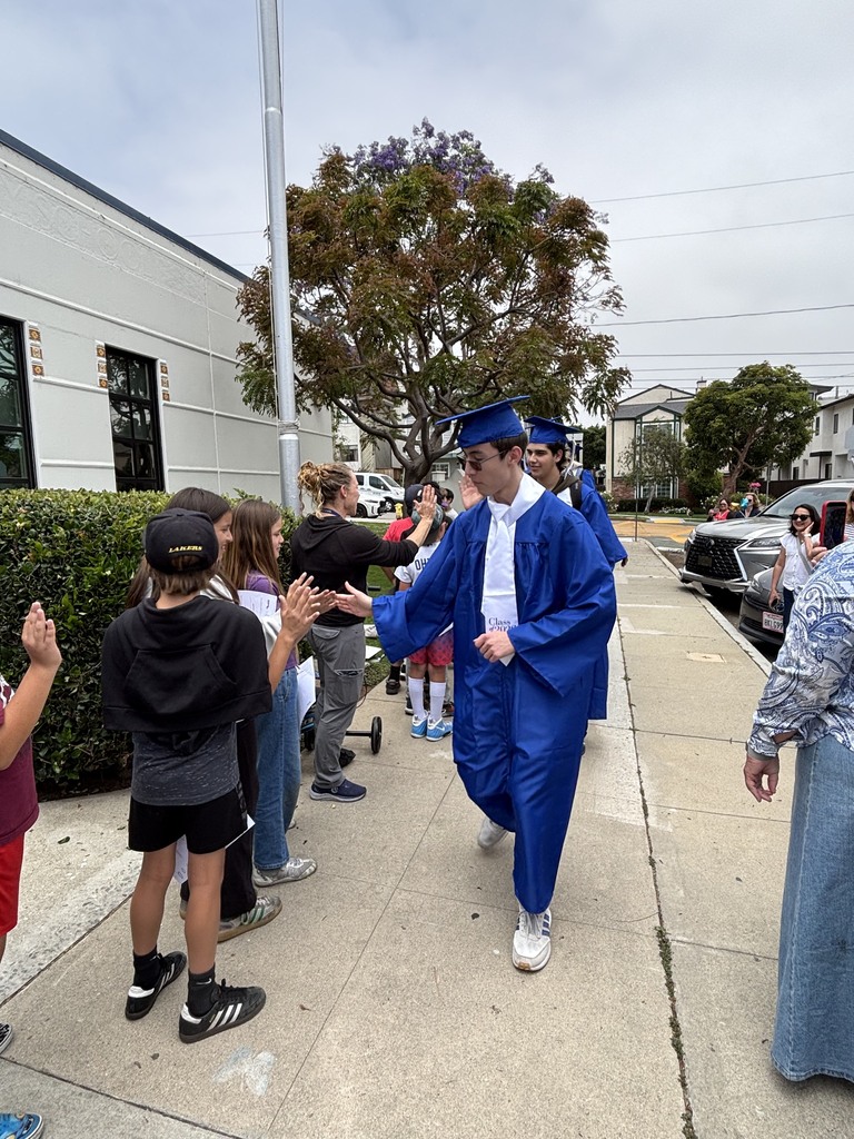 Graduating seniors in blue caps and gowns participate in a school walk, exchanging high-fives with younger students and families gathered outside.