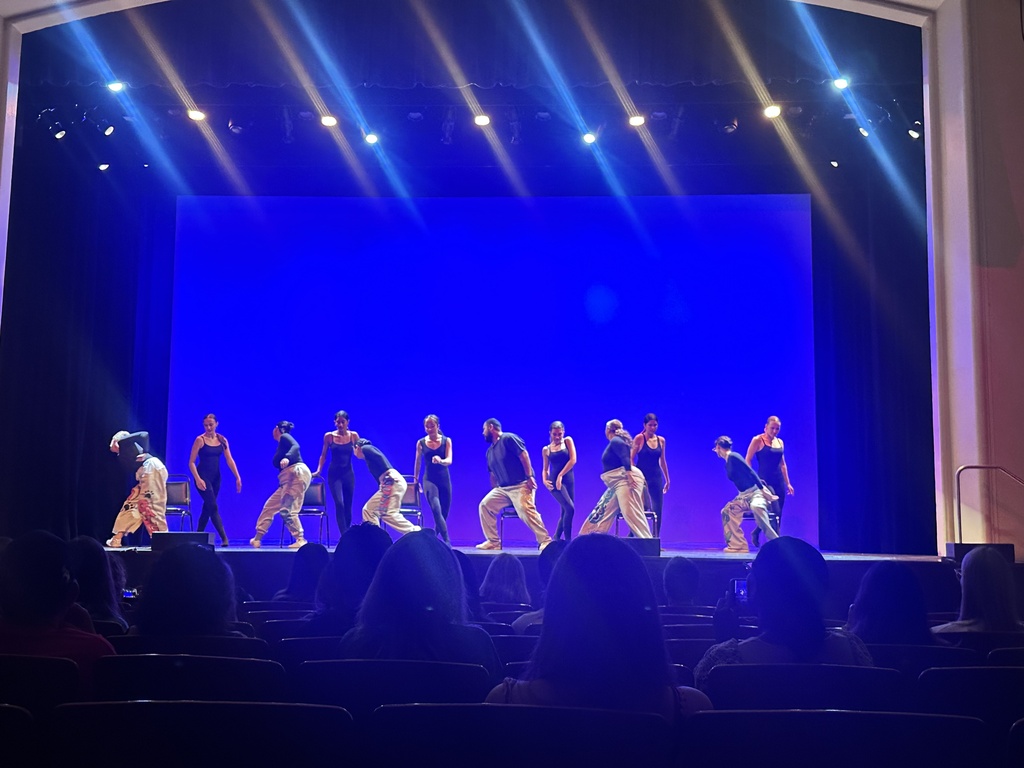 Students perform a choreographed dance on stage under blue lighting, with audience members watching from the theater seats.