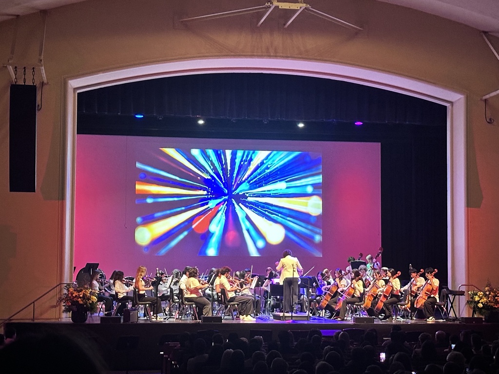 School orchestra performs before a packed audience as colorful abstract visuals are projected behind the ensemble.