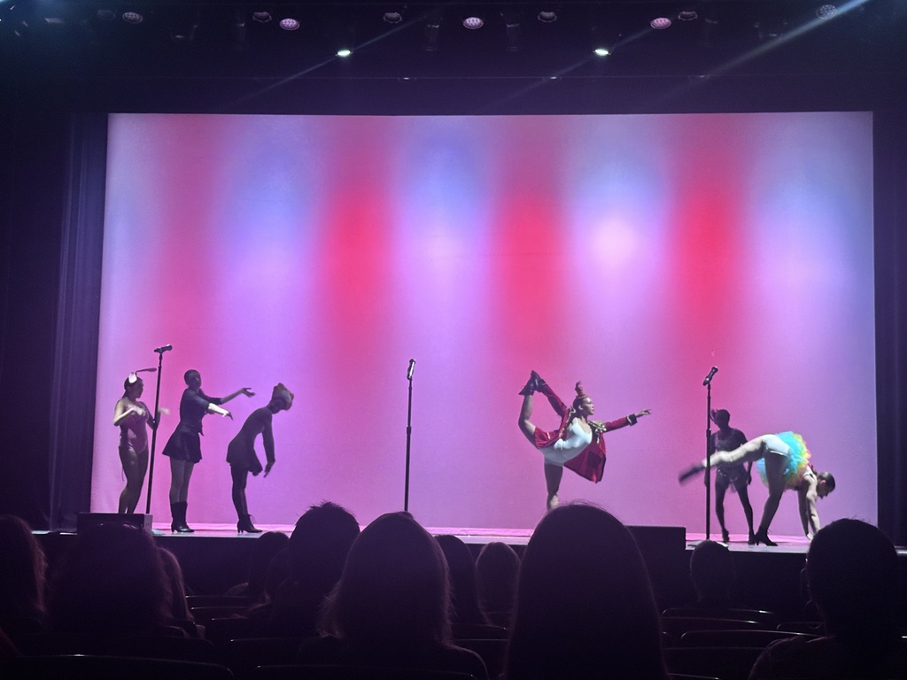 Performers in colorful costumes strike dramatic poses during a stage production illuminated by pink and purple lighting.