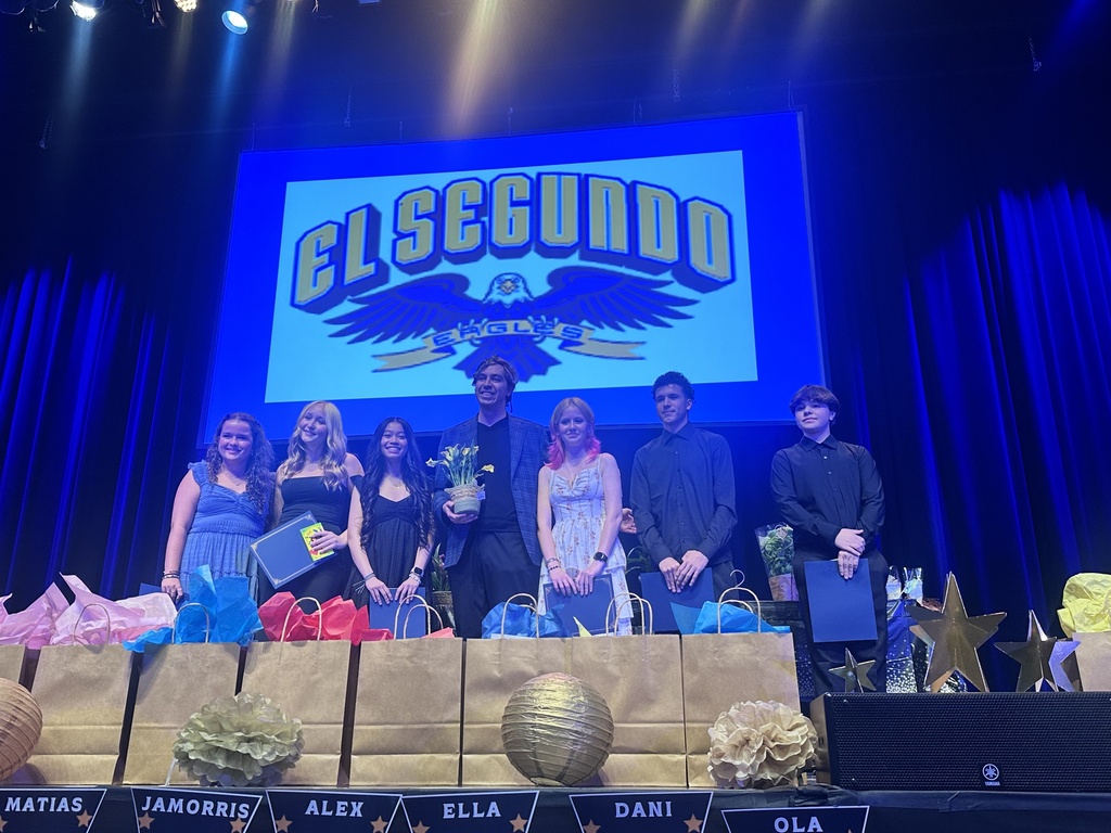Students and a teacher pose on stage with certificates and flowers during an academic awards presentation.