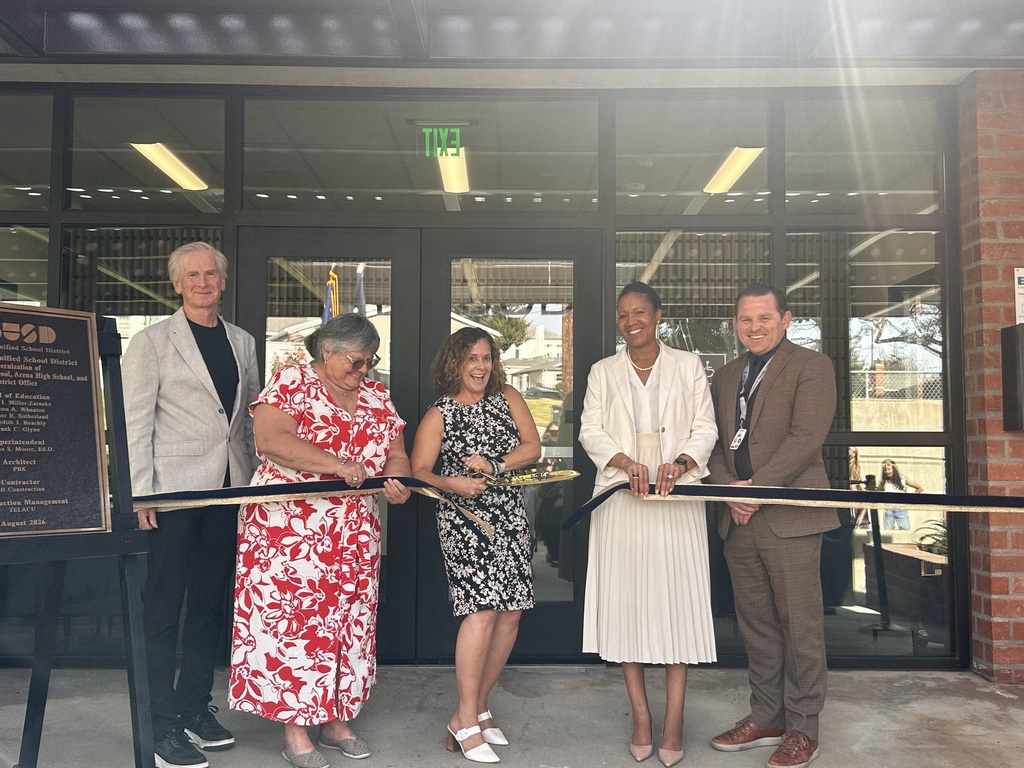 Five diverse individuals stand in front of glass doors, cutting a ceremonial ribbon to mark an official opening event.