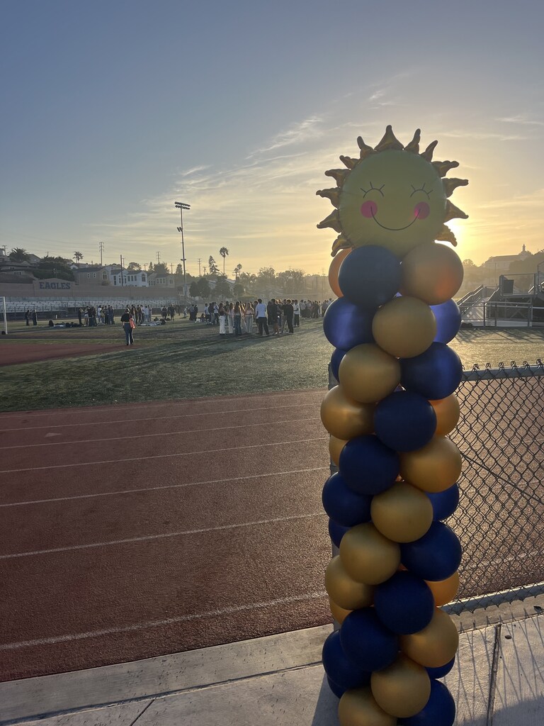 A blue-and-gold balloon column topped with a smiling sun decorates a track as students gather on the field at sunset.