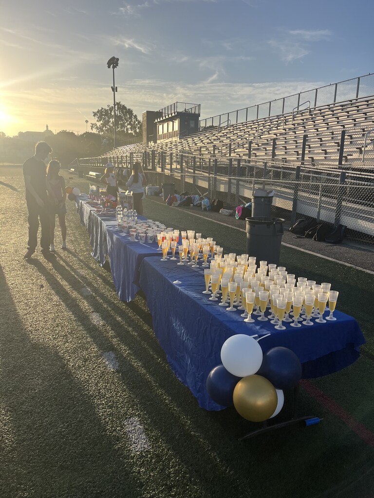 Blue-covered tables display rows of drinks and refreshments beside stadium bleachers in the warm sunset light.