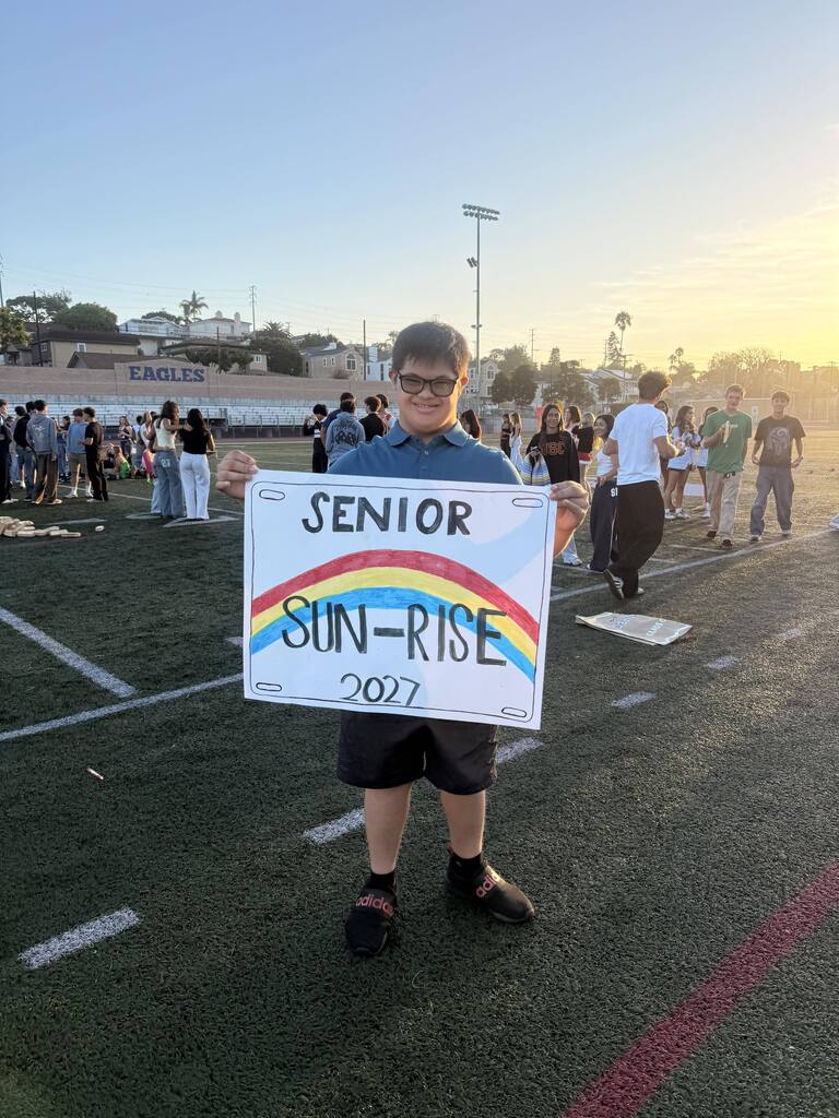 Smiling student holds a colorful “Senior Sun-Rise 2027” sign on the school field as the sun rises behind him.