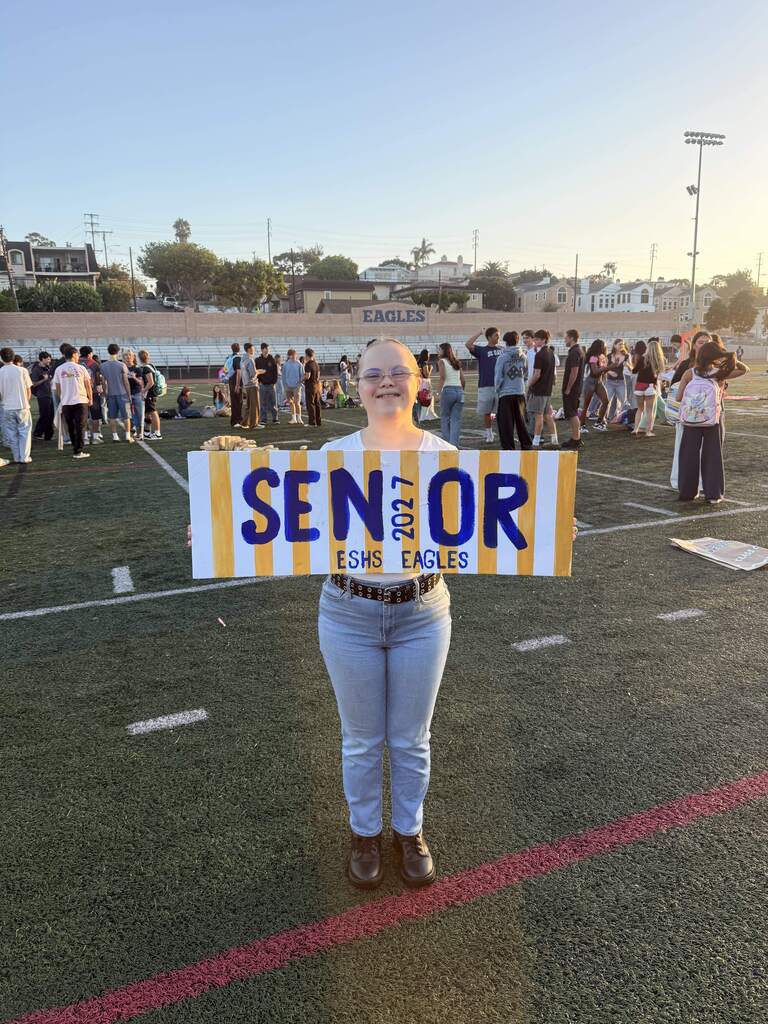 Smiling student holds a handmade “Senior 2027 ESHS Eagles” sign on the school field during a senior gathering.
