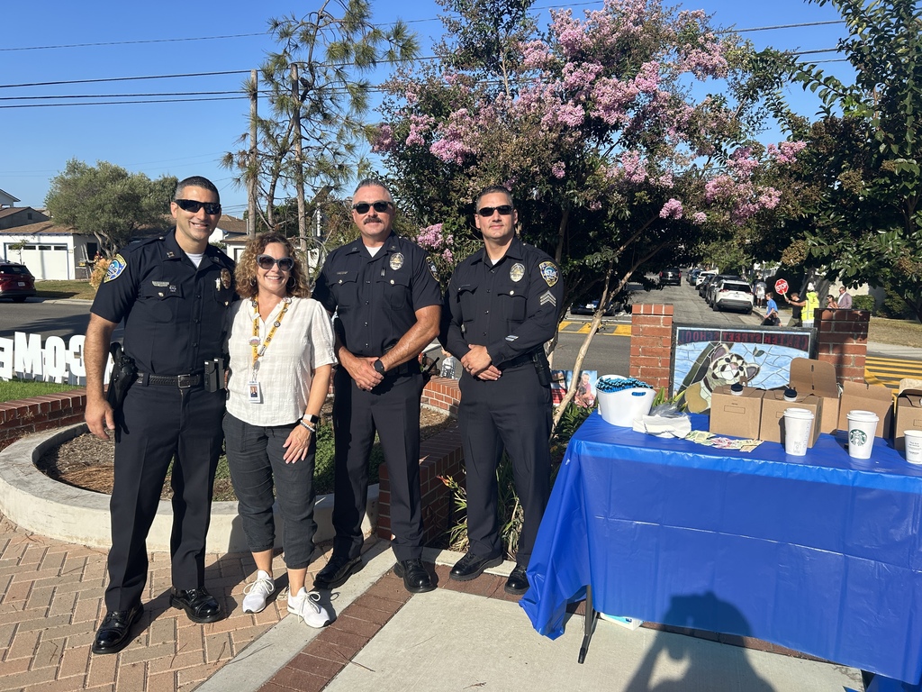 Three police officers and a woman stand together outdoors near a table with a blue tablecloth and boxes under a blooming tree.