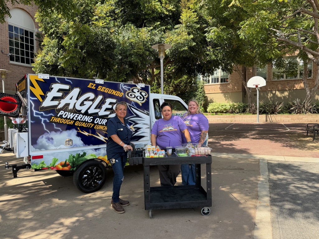 Three school nutrition staff members pose beside an El Segundo Eagles mobile lunch cart stocked with food and drinks.