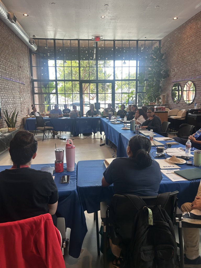 A group of people seated around a large U-shaped table covered with blue tablecloths in a bright room with large windows and exposed brick walls.
