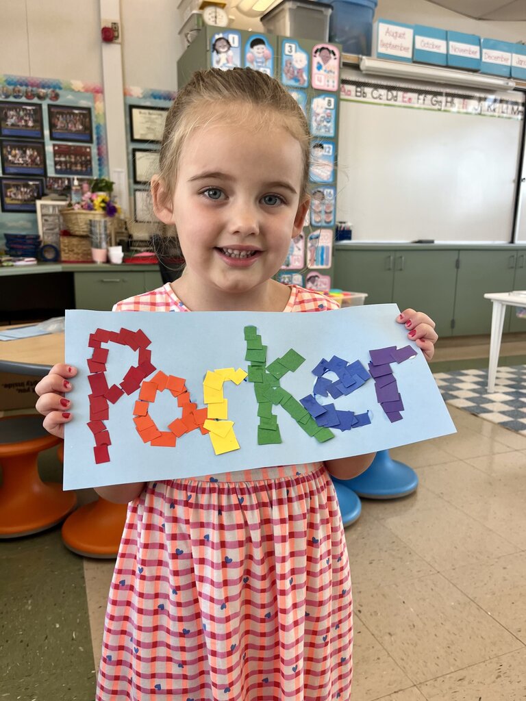 Young girl in a classroom holding a colorful paper craft spelling 'Parker' with torn paper pieces.