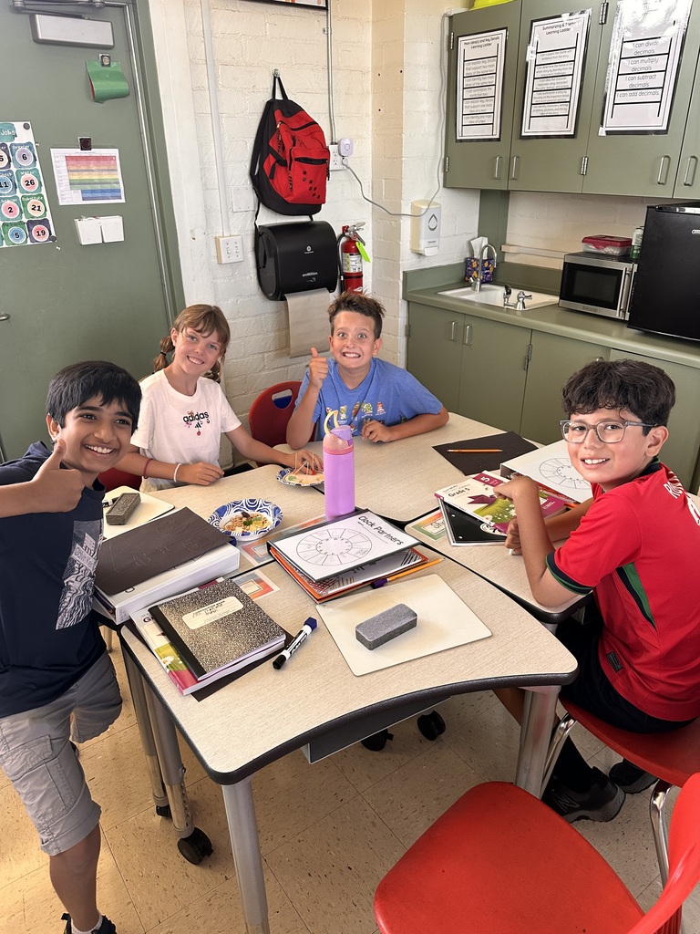 Four smiling students sitting around a classroom table with notebooks, papers, and a water bottle, engaged in group work.