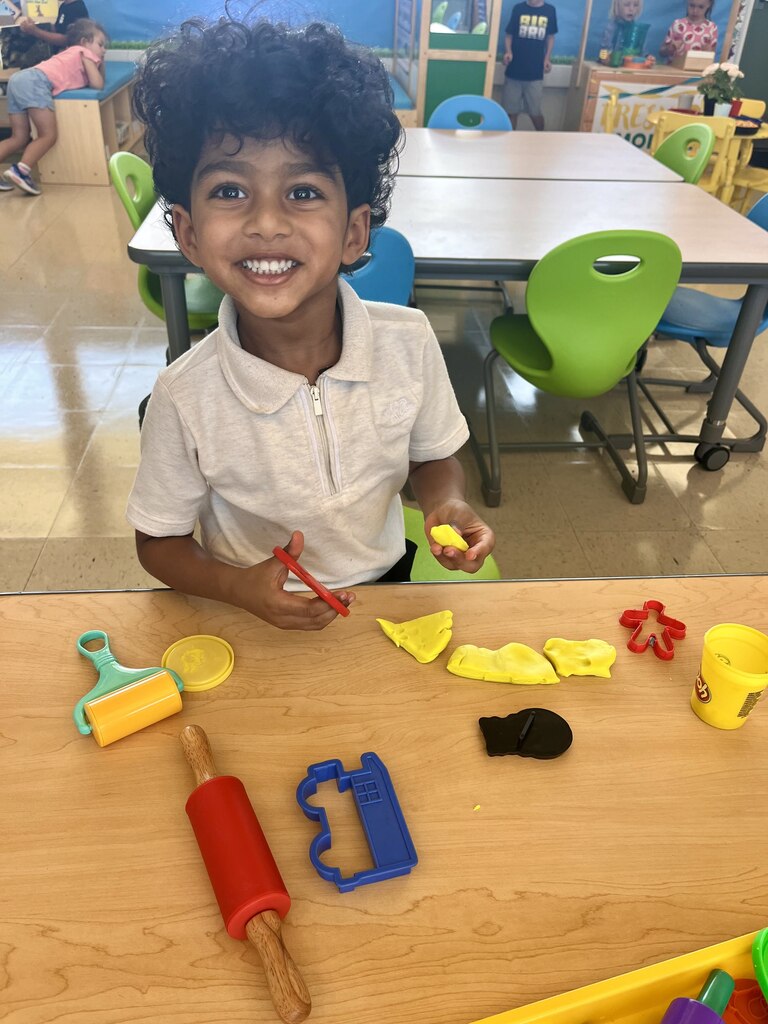 Smiling young boy with curly hair playing with yellow playdough and various shaping tools at a classroom table.