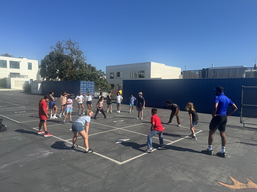 Students and an adult gather in a circle on a sunny school playground while participating in an outdoor activity.