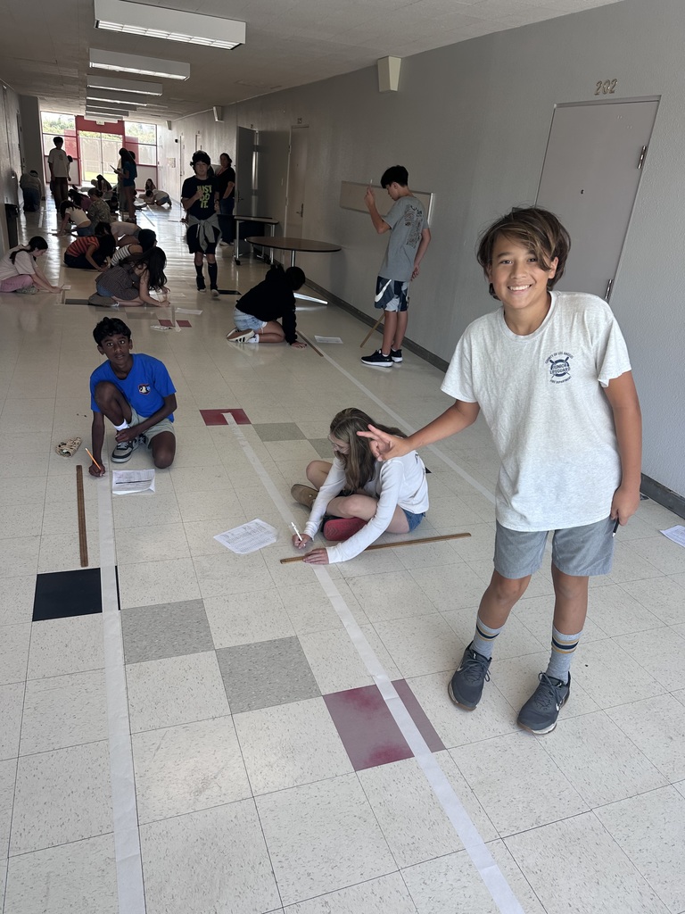 Children engaged in a hallway activity measuring and marking the floor with tape and paper, with one boy smiling and posing in the foreground.