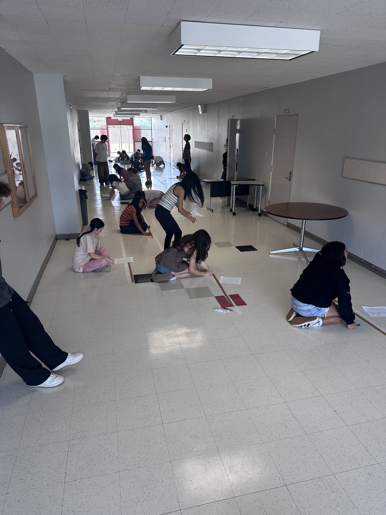 Children spread out on a school hallway floor, engaged in drawing or writing activities on large sheets of paper.