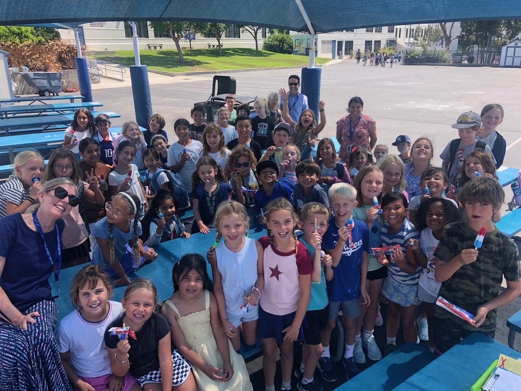 A large group of smiling elementary school children and teachers gathered under a shaded picnic area enjoying popsicles on a sunny day.