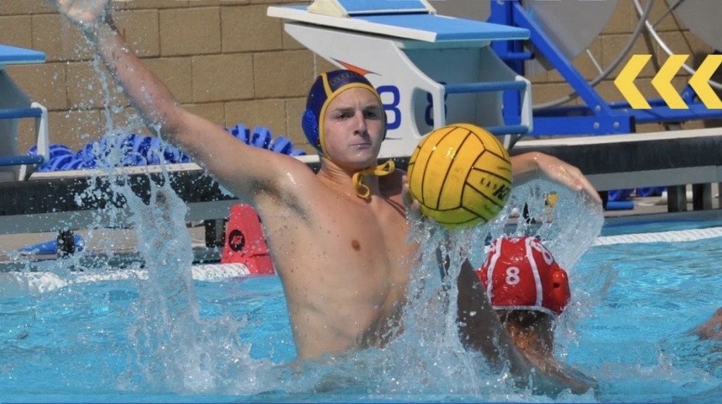 Water polo player in a yellow cap preparing to throw the ball while an opponent in a red cap tries to block him in the pool.