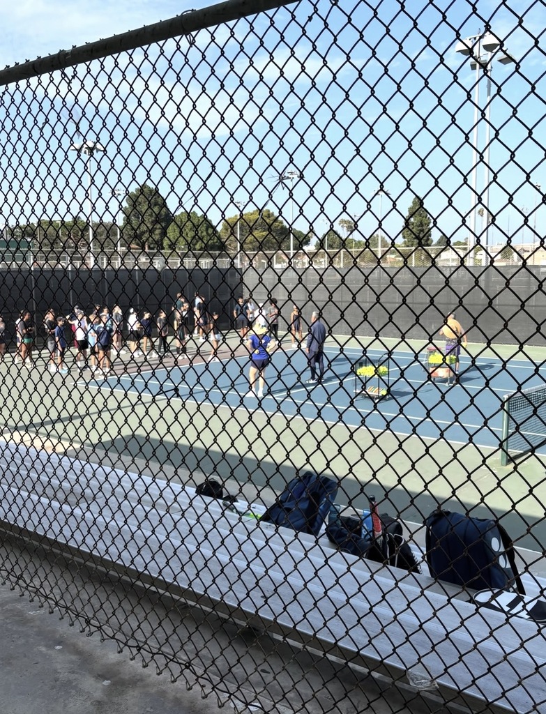 View through a chain-link fence of a busy outdoor tennis court with players practicing and equipment bags on the sidelines.