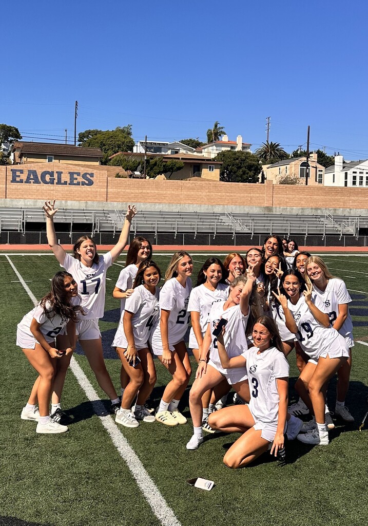 A group of student athletes in white uniforms pose playfully together on a sunny school athletic field.