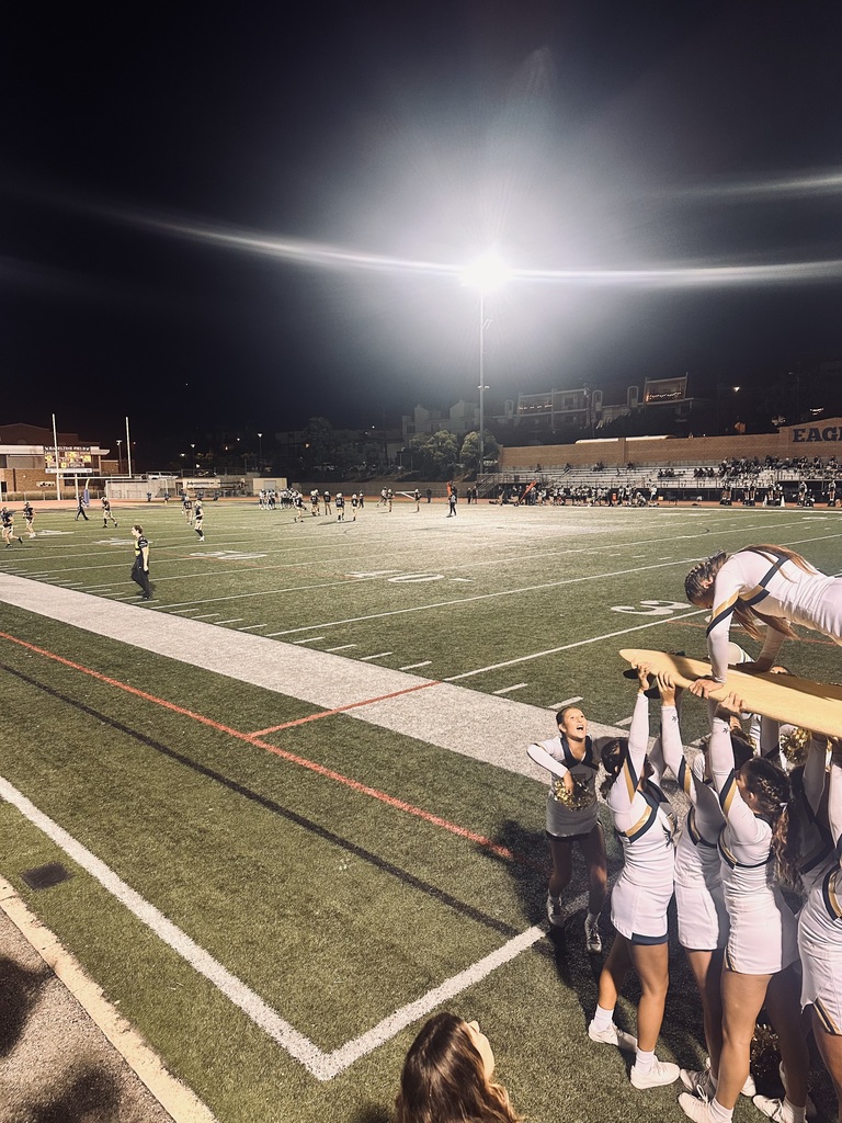 Cheerleaders perform a stunt along the sideline during a nighttime football game under stadium lights.