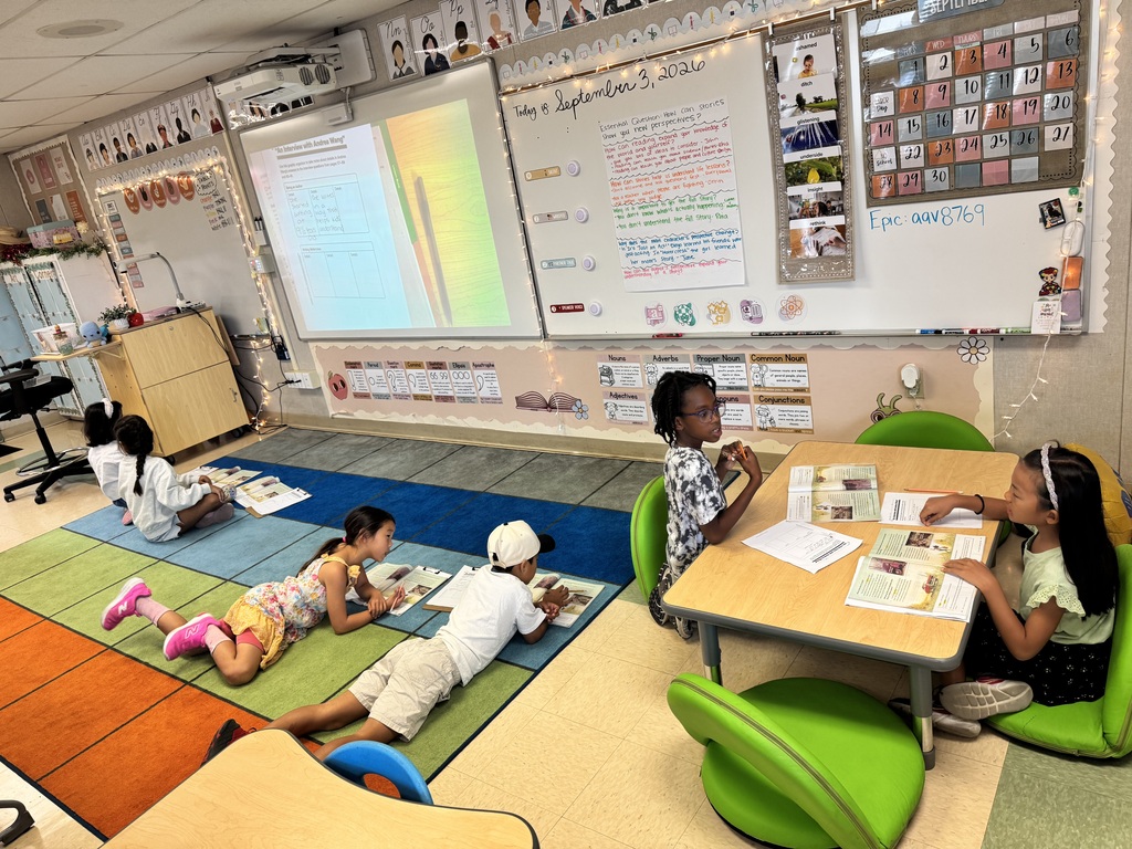 Four children engaged in reading activities in a colorful classroom with educational posters and a whiteboard displaying notes and a calendar.