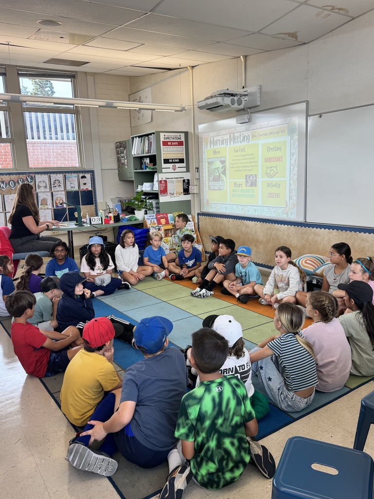 Elementary students sitting in a circle on a colorful classroom rug during a group lesson with a teacher at the front.