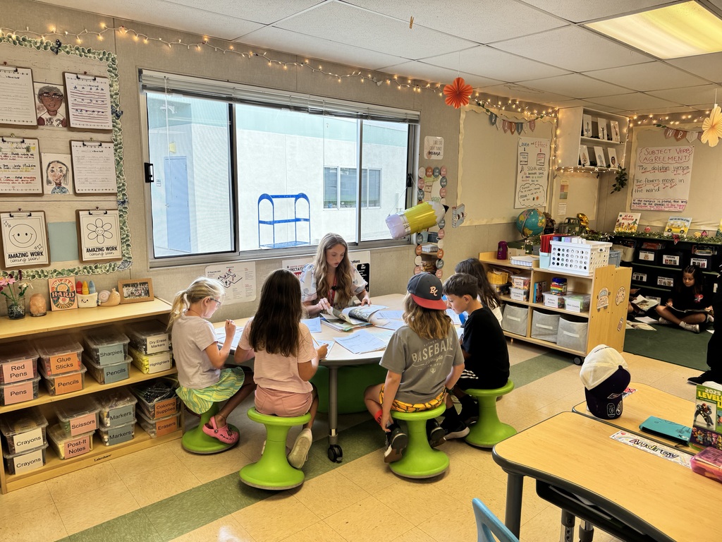 A teacher reads a book to five young students seated on green stools around a low table in a colorful classroom.