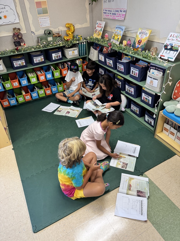 Four children sitting on a green mat in a classroom, reading books and worksheets spread out around them.