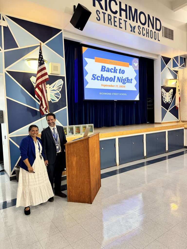 Two school staff members stand near a podium in a decorated auditorium with a 'Back to School Night' presentation on screen.