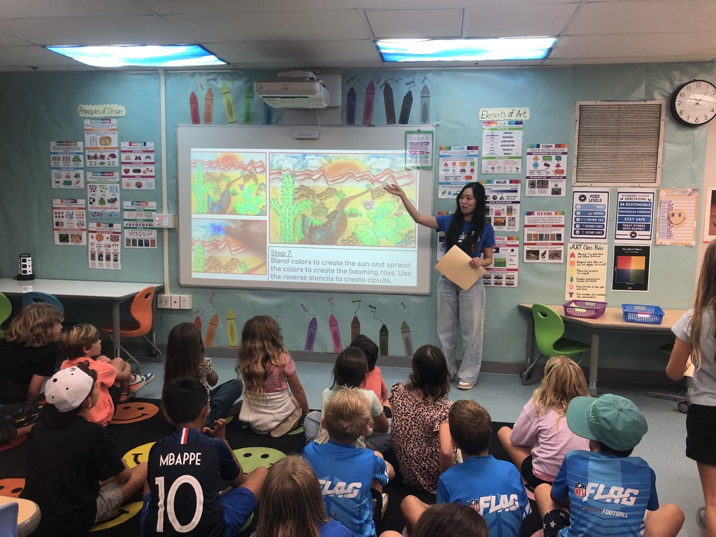 Teacher explaining a colorful art project to a group of attentive young students seated on the floor in a bright classroom.