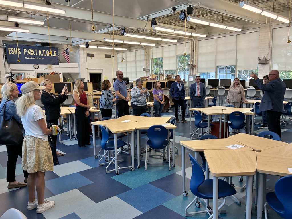 Group of adults standing in a classroom with hexagonal tables and blue chairs, listening to a speaker near the windows.