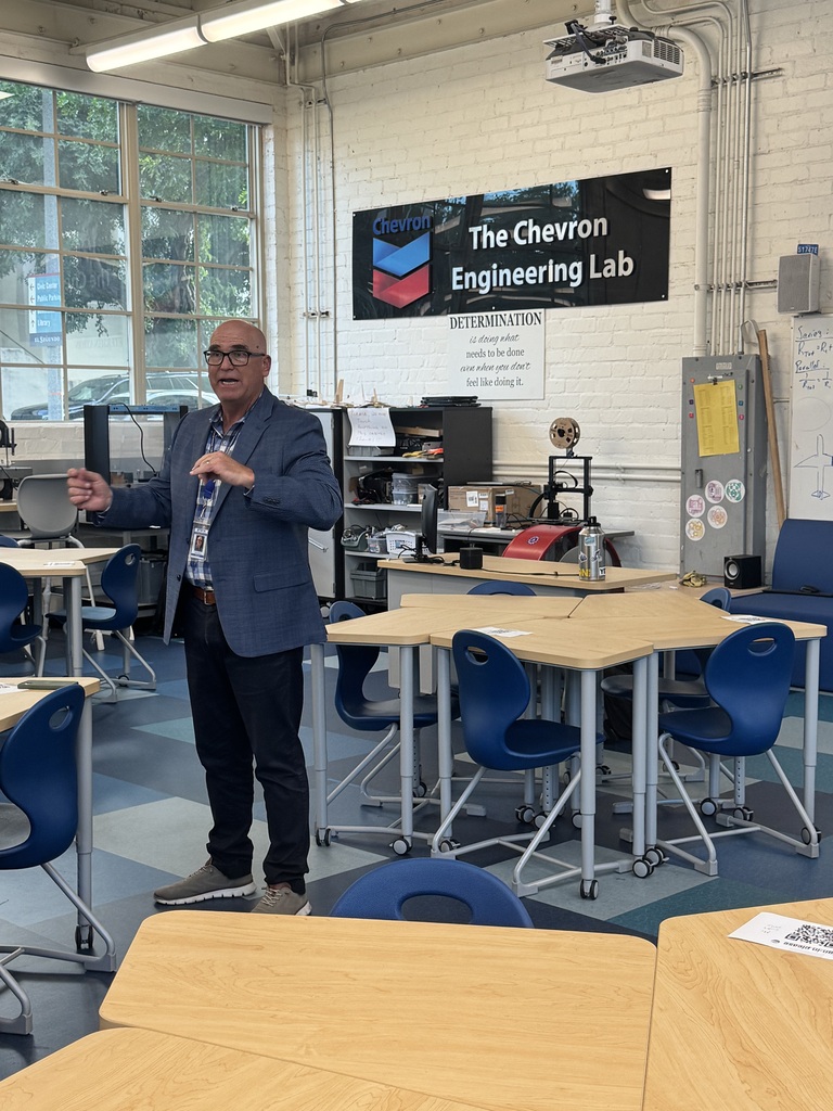 A man in a blazer speaking inside The Chevron Engineering Lab with blue chairs and wooden tables around.