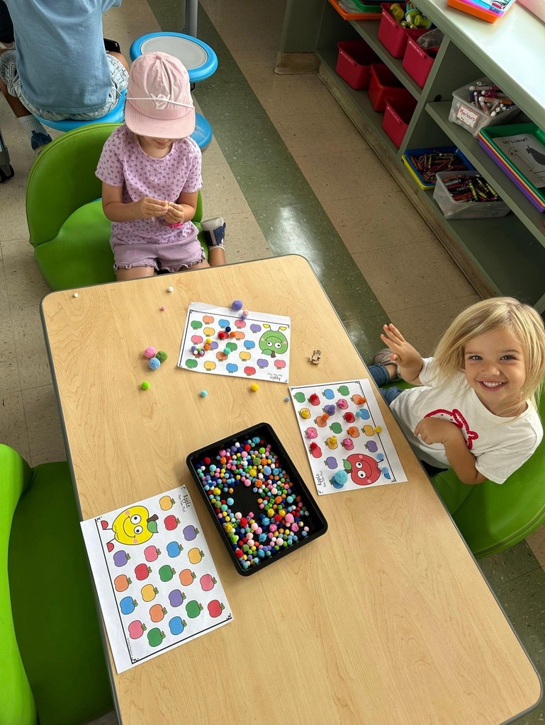 Two children use colorful pom-poms to complete apple-themed matching and sorting activity sheets at a table.