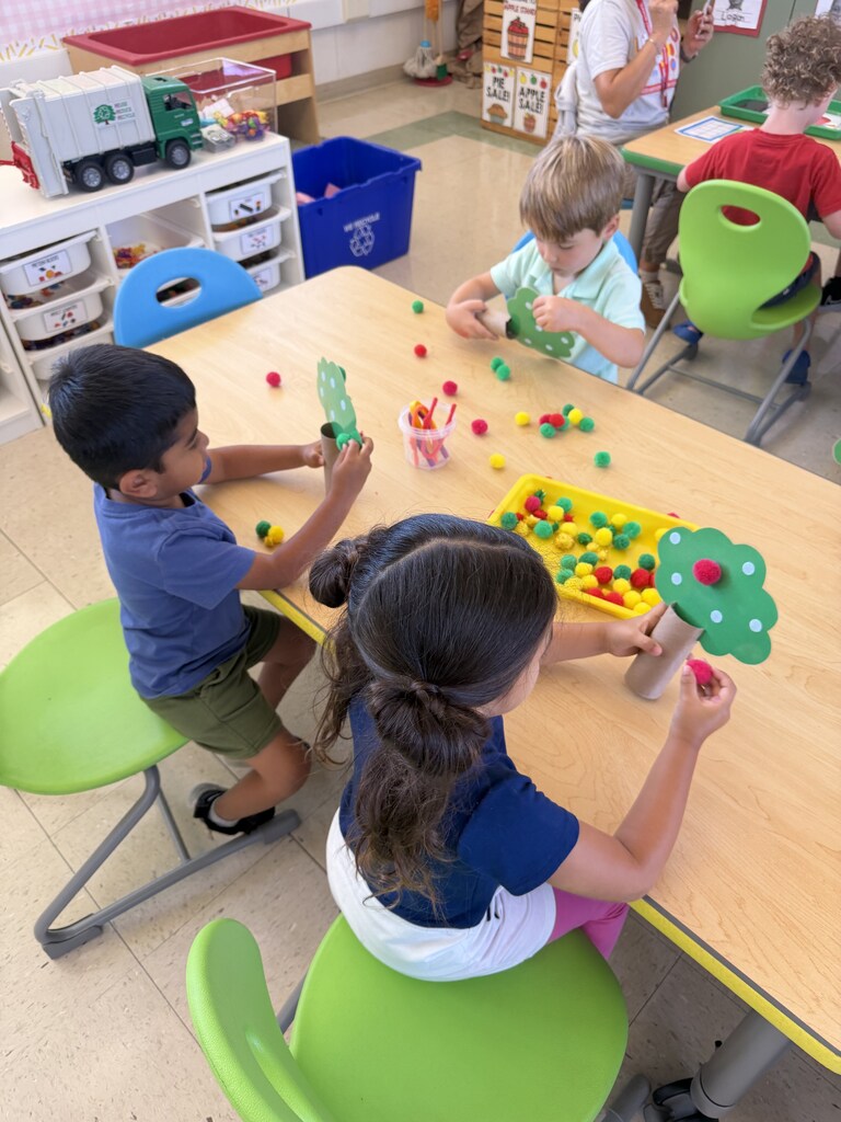 Three children create paper trees with colorful pom-pom apples during a hands-on classroom activity.