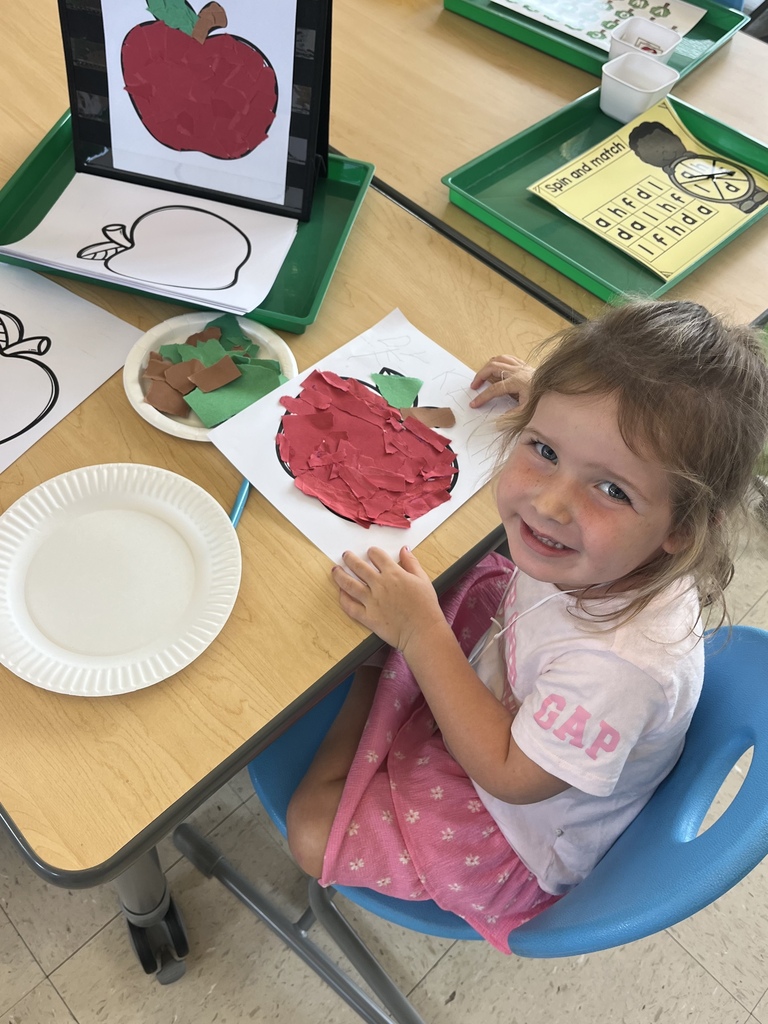 A smiling child displays a red paper apple collage made with torn paper during an apple-themed classroom activity.