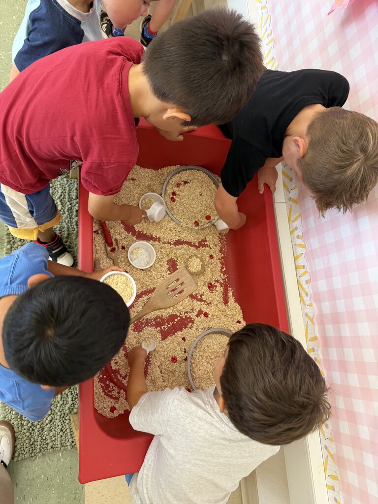 Children scoop and pour oats with bowls and utensils while exploring an apple-themed classroom sensory bin.